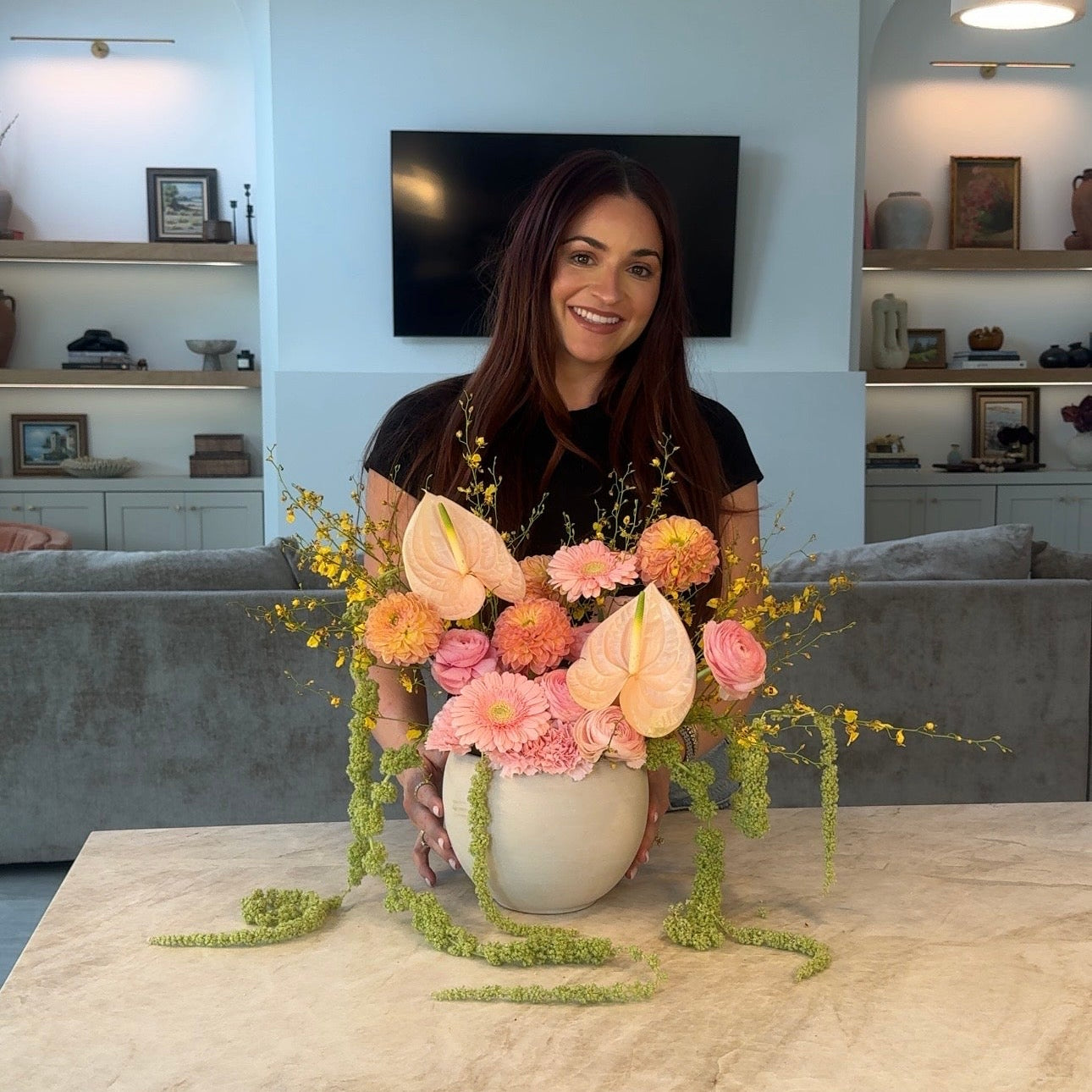 A smiling woman stands behind a floral arrangement of pink, peach, and orange flowers with green trailing vines in a white vase, set on a light marble table in a modern living room.