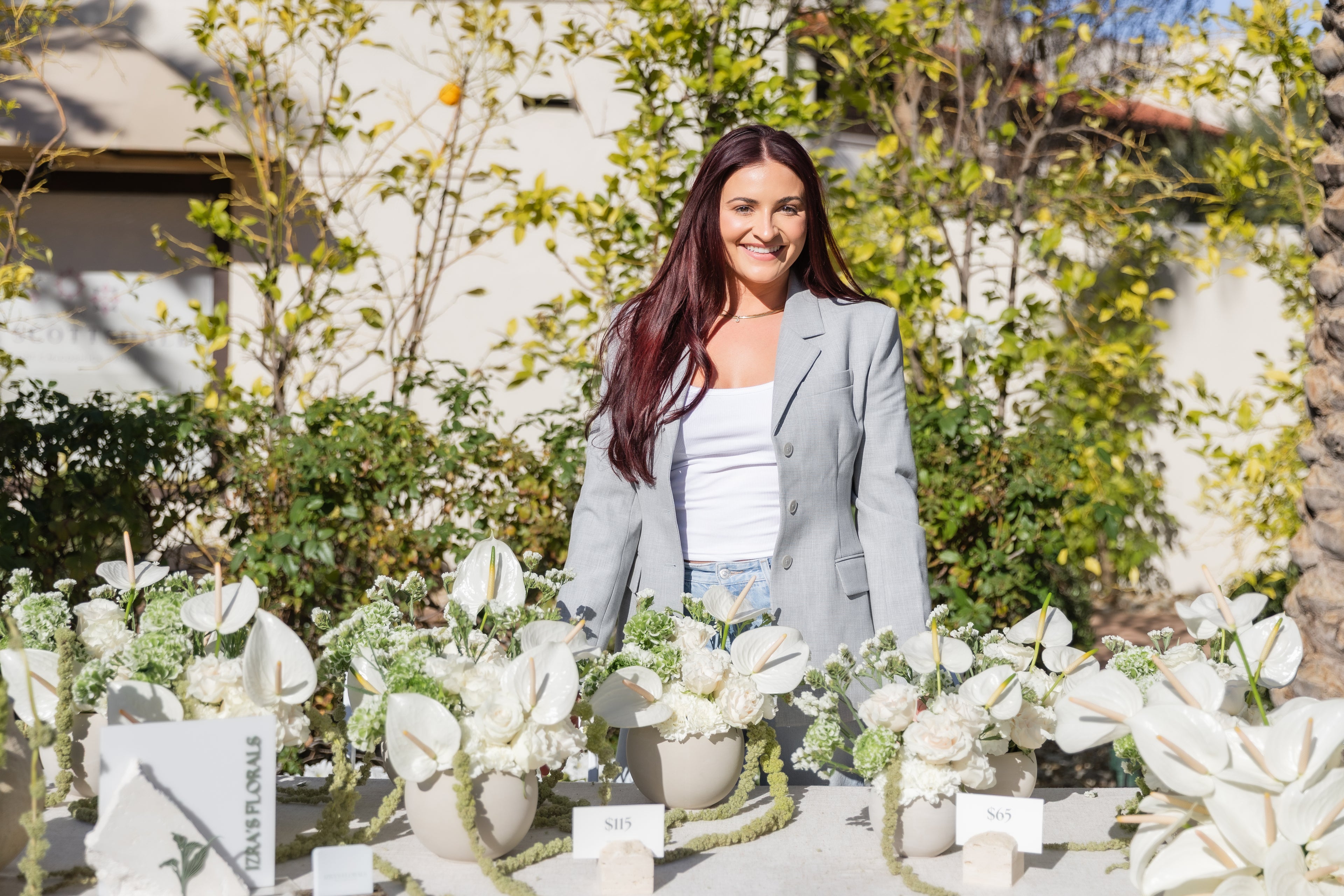 A woman with long dark hair stands behind a table decorated with white flowers and greenery, wearing a light gray blazer over a white top. The setting is outdoors with trees and a building in the background.